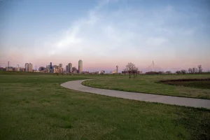 A wide-angle skyline view of Dallas, Texas, captured from across a vast green field at dusk under a pale, pastel sky. A paved walking trail winds through the grass in the foreground, curving towards the city, softly lit by the evening light.